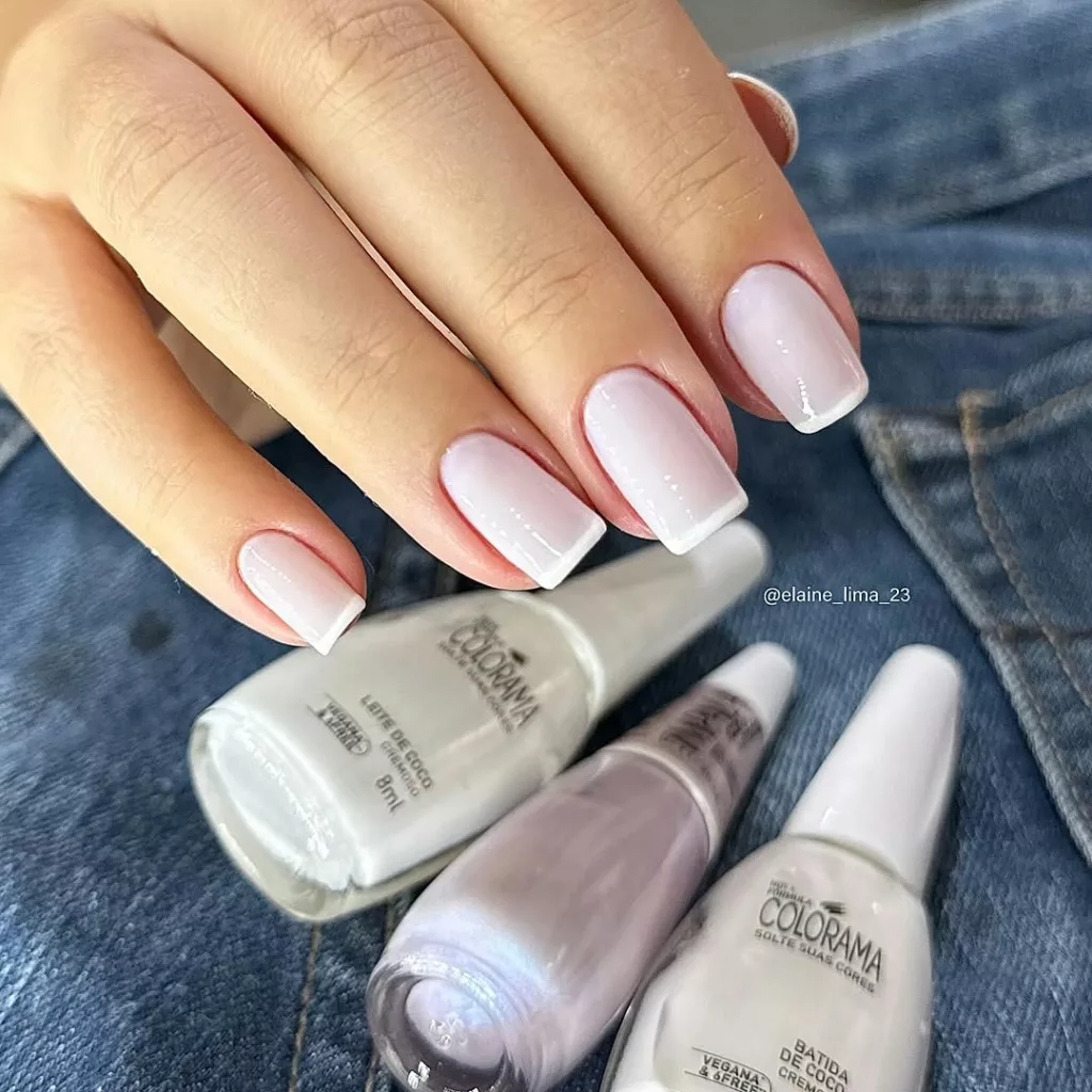 Unhas com francesinha clássica em base leitosa e acabamento brilhante, no formato quadrado curto, usando esmaltes brancos e cintilantes da Colorama sobre jeans.