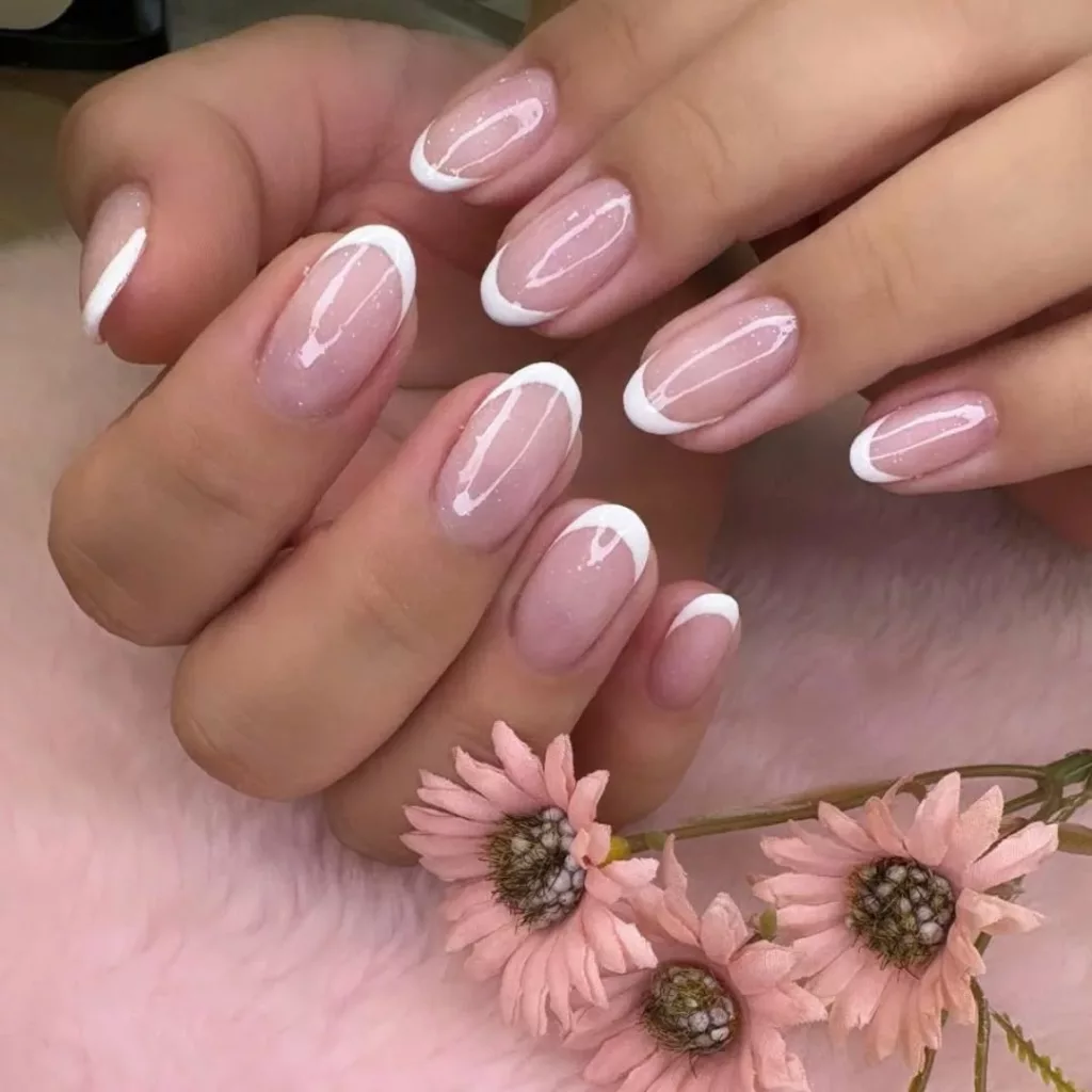 Francesinha oval com base rosada e ponta branca arredondada, em unhas curtas e delicadas, acompanhadas de flores artificiais cor-de-rosa.