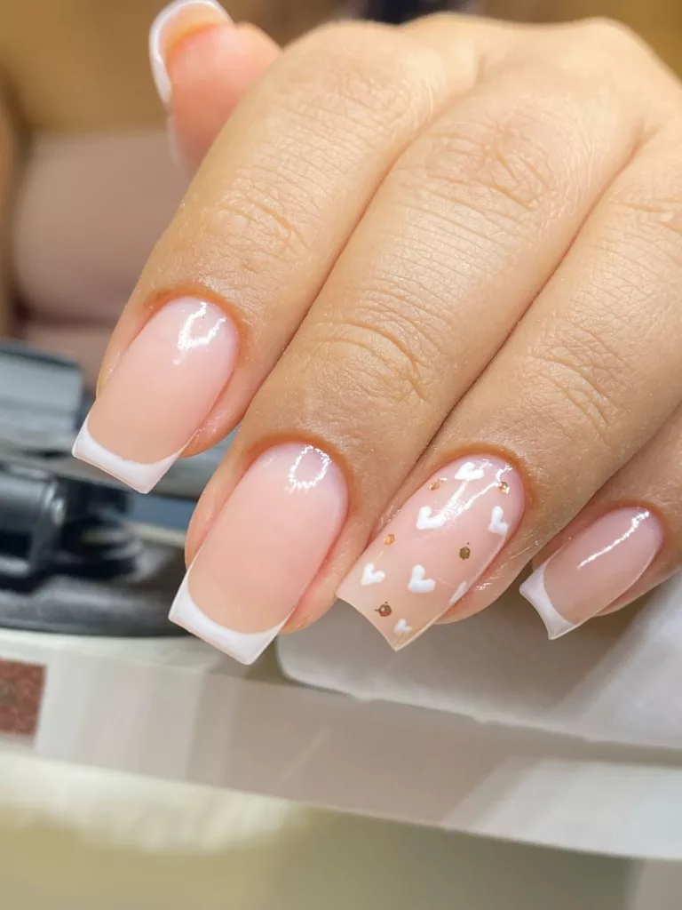 Unhas quadradas com francesinha branca tradicional e destaque para uma unha decorada com mini corações brancos e detalhes dourados.