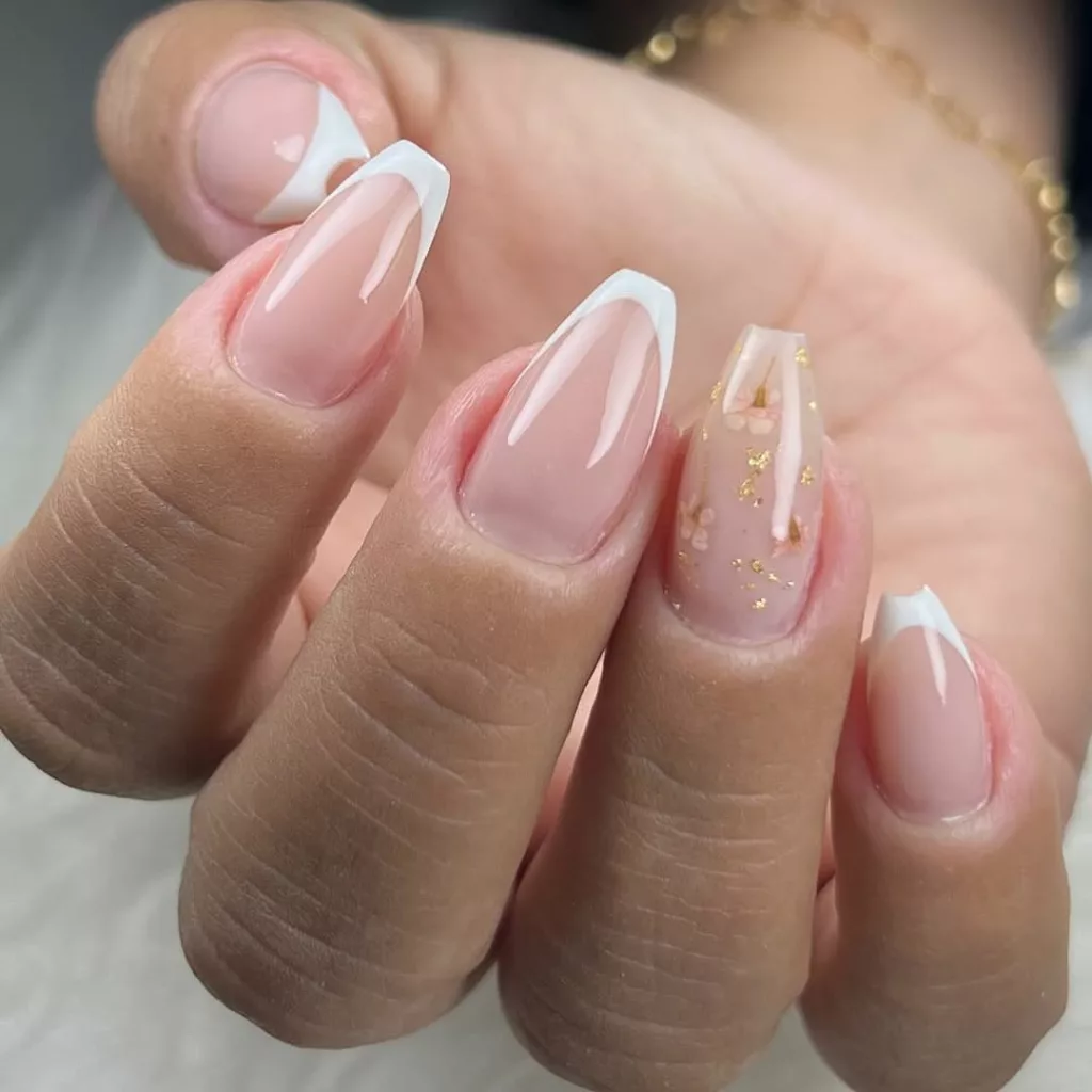 Francesinha moderna com traço curvo branco em unhas de formato amendoado, com uma unha decorada com flocos dourados e flores encapsuladas.