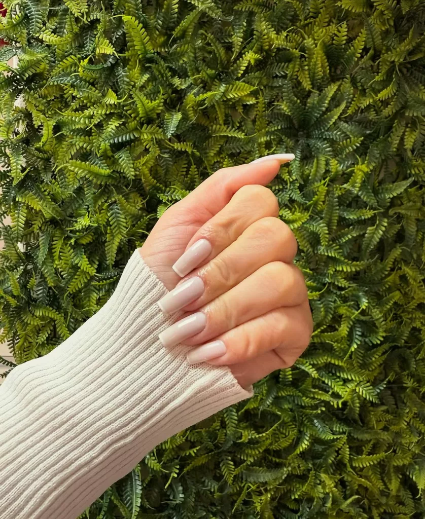 Francesinha nude fosca em unhas quadradas longas, com acabamento acetinado e fundo acinzentado sobre fundo de folhagens verdes.
