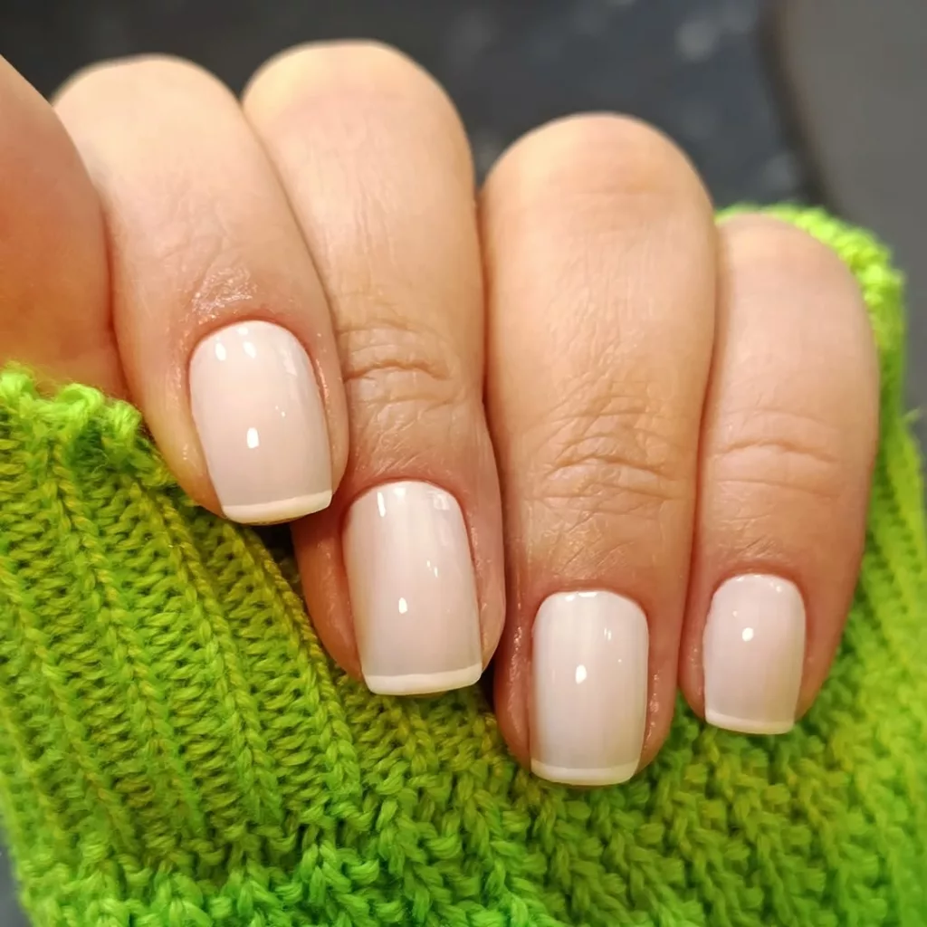 Francesinha fininha e discreta em unhas curtas com acabamento translúcido nude, combinando com uma blusa de lã verde vibrante.