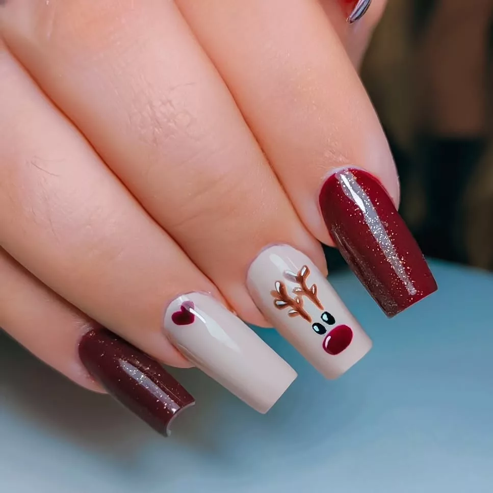 Nail art de Natal com esmalte vinho, base nude e desenho de rena com nariz vermelho, criando uma proposta divertida e temática para as festas.