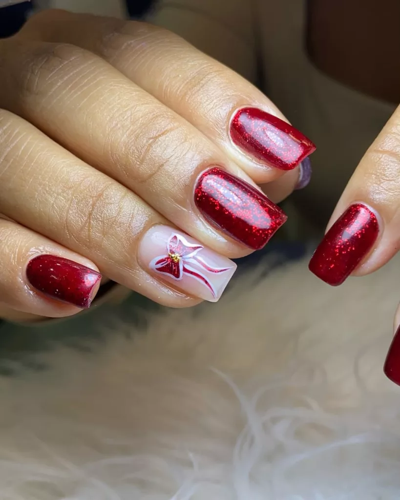 Unhas vermelhas com glitter e desenho de laço natalino branco e vermelho, destacando o espírito de presente e celebração da época.