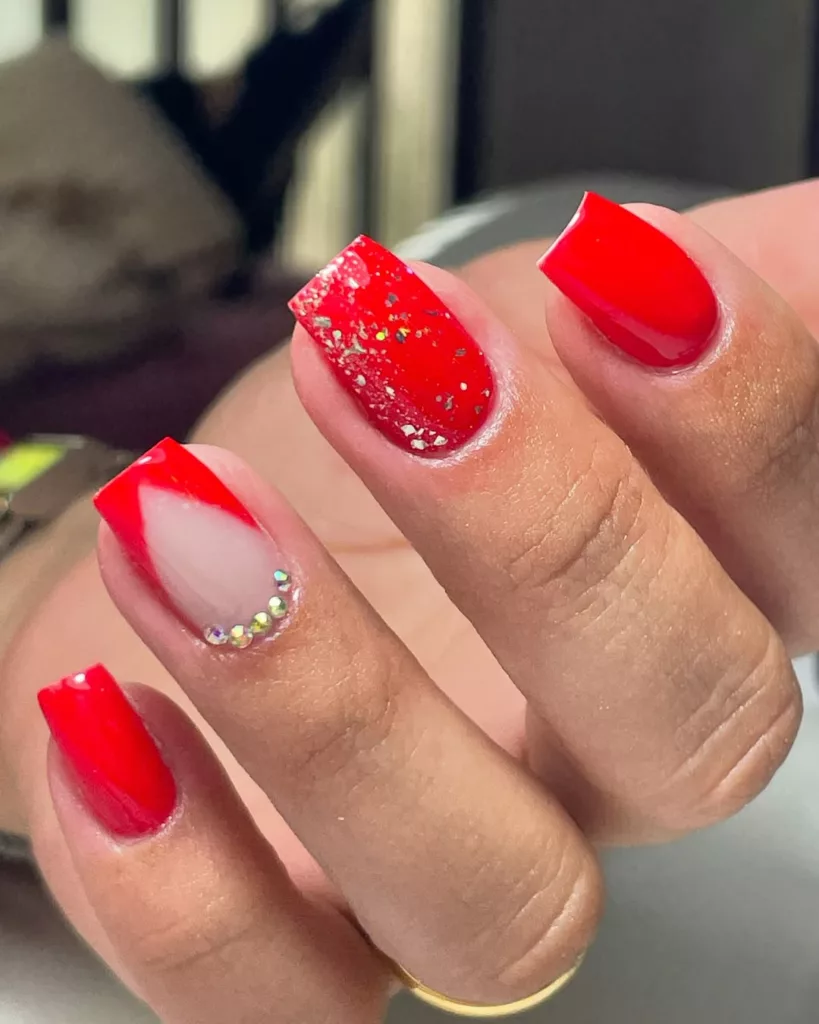 Nail art para o Natal com esmalte vermelho, glitter prateado e aplicação de pedrinhas na base da unha, criando um efeito sofisticado e festivo.