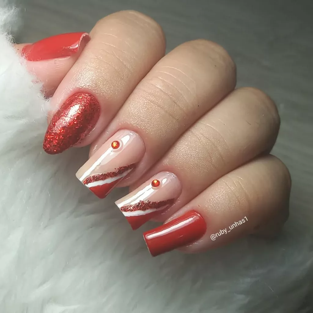 Nail art com glitter vermelho, linhas diagonais em branco e vermelho e aplicação de pedrinhas brilhantes, criando uma composição elegante e moderna.