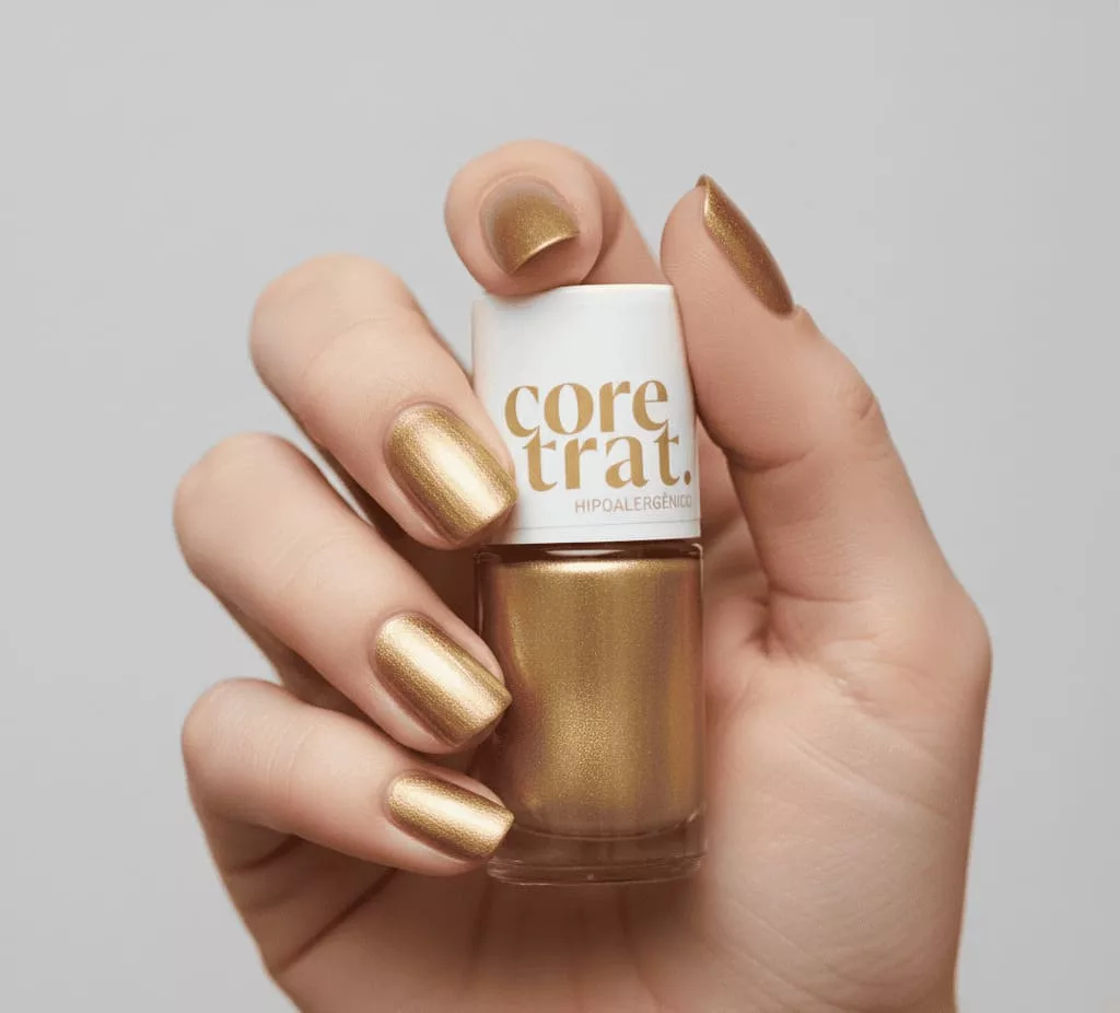Unhas com esmalte dourado metalizado de acabamento uniforme e brilho intenso, segurando frasco do esmalte Coretrat Metálico Blant Dourad, ideal para um visual luxuoso e sofisticado no Natal.