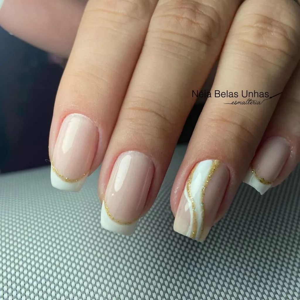 Unhas curtas com francesinha branca e detalhe dourado, incluindo um dedo com desenho curvado delicado, ideal para um visual minimalista na virada.
