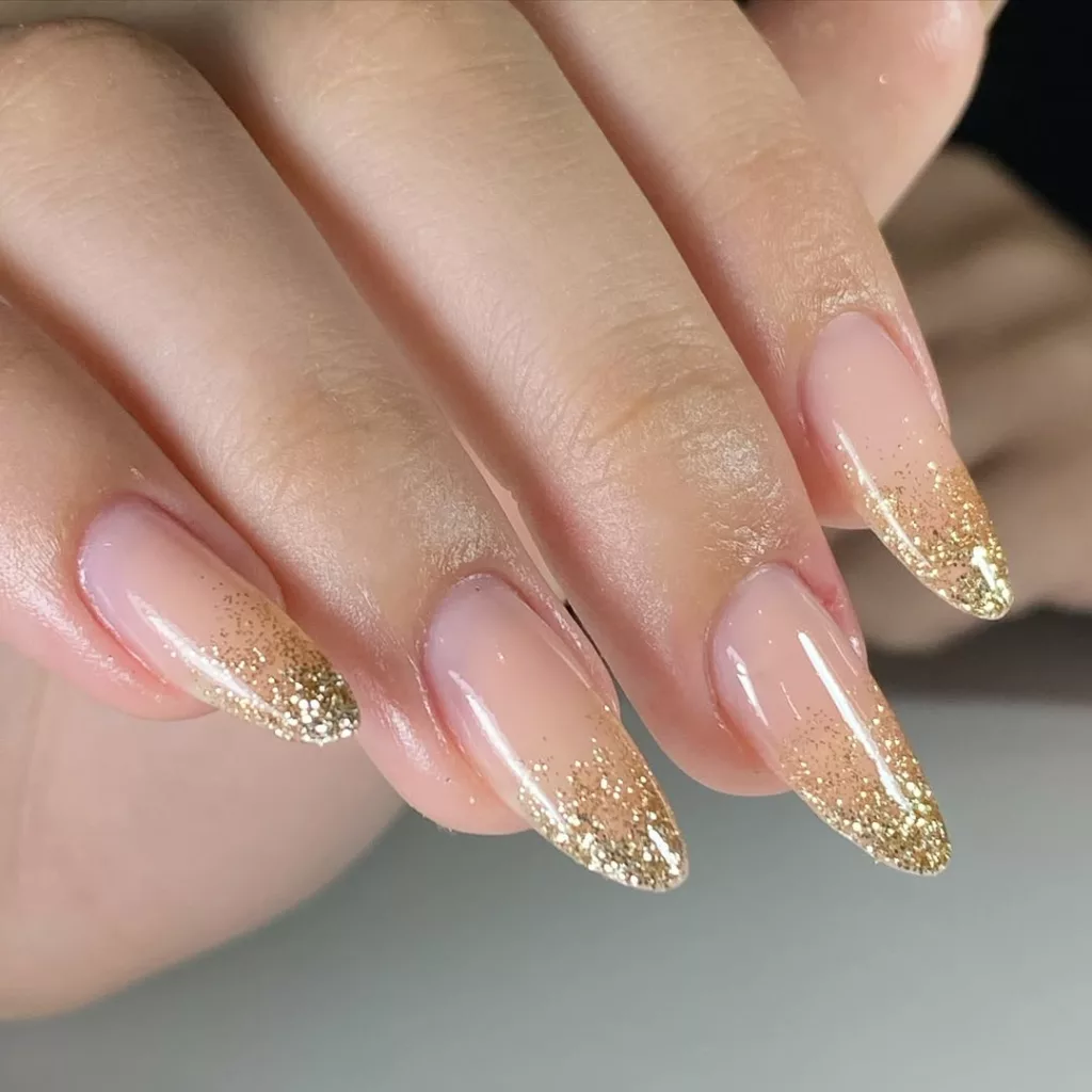 Unhas amendoadas nude com glitter dourado esfumado nas pontas, formando um degradê elegante e festivo.