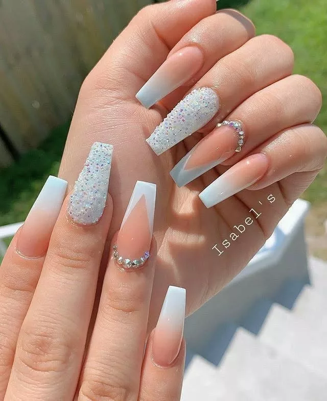 Unhas longas com degradê branco, dedo com glitter iridescente e aplicação de pedrinhas, resultando em um design festivo e marcante.