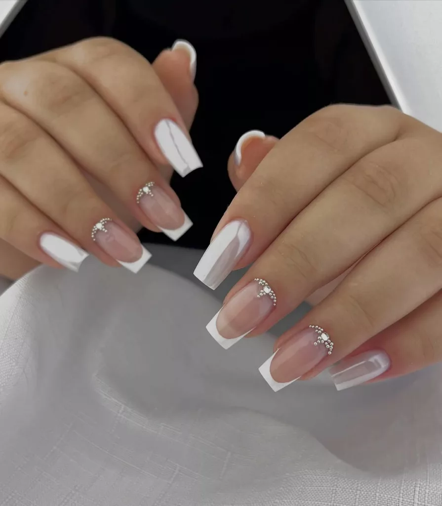 Unhas com francesinha milky e aplicação de pedrinhas no arco da cutícula, criando um visual delicado e elegante.