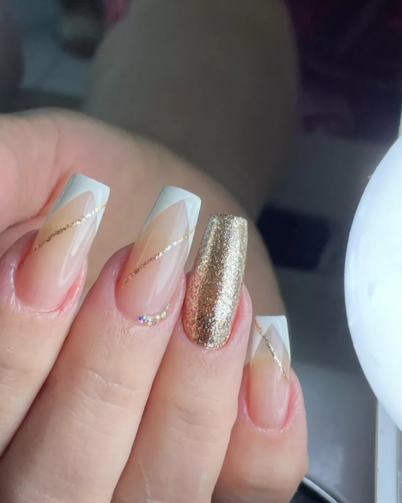 Unhas nude com traços diagonais dourados e um dedo totalmente dourado em glitter, combinando modernidade e brilho para a virada do ano.
